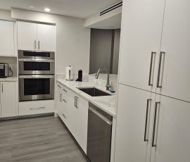 Right-side view of a custom U-shaped kitchen in Hillsboro Beach, Florida, featuring OneSkin Sunny White Super Matte flush-panel cabinetry, a tall pantry cabinet, kitchen sink, breakfast bar, Tuscani Quartz countertops, and built-in double wall ovens.