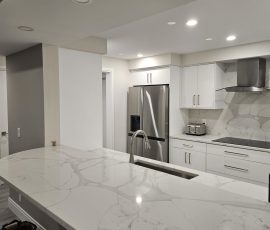 Custom U-shaped kitchen, Hillsboro Beach, Florida, viewed from the dining room, featuring a curved Tuscani Quartz breakfast bar pass-through, OneSkin Sunny White Super Matte flush-panel cabinetry, stainless steel refrigerator, cooktop with stainless steel range hood, and integrated LED accent lighting in the soffit and kitchen.